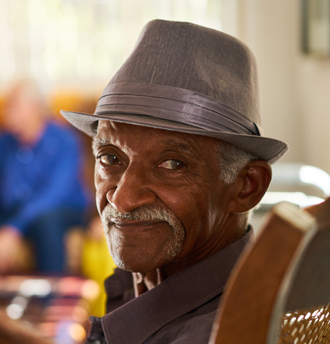 Portrait,Of,Elderly,Black,Man,Looking,At,Camera,In,Retirement Portrait of elderly black man looking at camera in retirement home, with group of friends in background. Patients relaxing in hospice for seniors.
